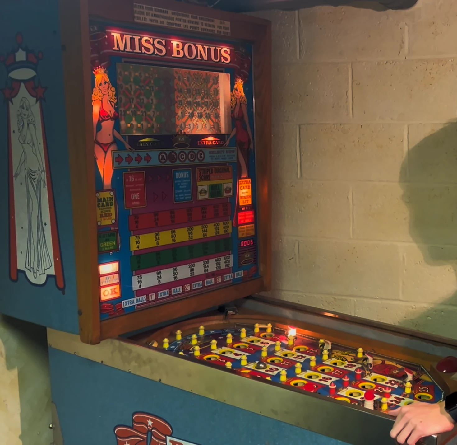 The WIMI Miss Bonus bingo pinball machine in a basement, with its lit Miss Bonus backglass and the numbered playfield below it